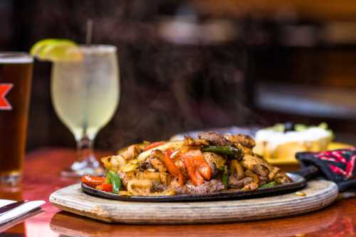 Sizzling fajitas with grilled meat and vegetables on a wooden platter, accompanied by a beer and a cocktail.