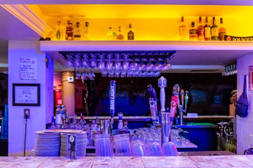 A well-stocked bar with illuminated shelves, various liquor bottles, and hanging glasses against a colorful backdrop.