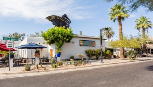 A charming restaurant with outdoor seating, surrounded by palm trees and desert landscaping, under a clear blue sky.