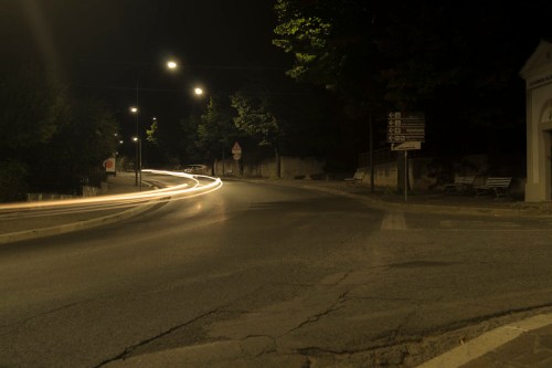 A quiet, dimly lit road at night with light trails from passing vehicles and trees lining the street.