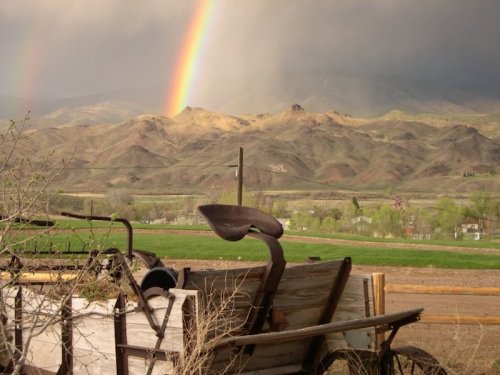 A rustic plow sits in the foreground with mountains and a vibrant rainbow in the background under a cloudy sky.