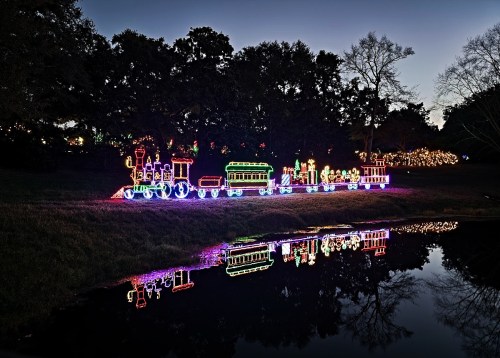 A colorful illuminated train display reflects in a calm pond at dusk, surrounded by trees and festive lights.