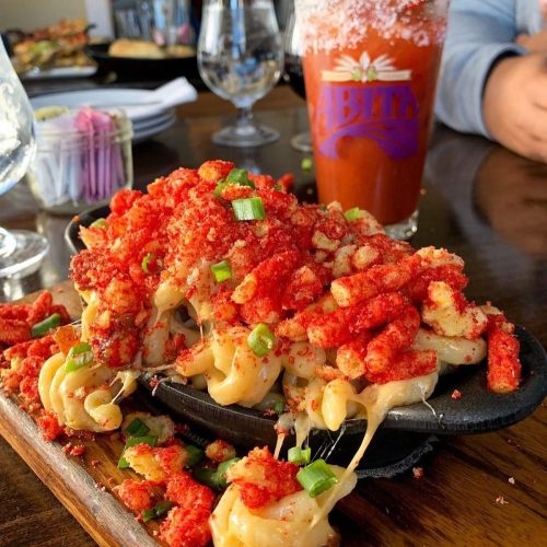 A sizzling plate of cheesy pasta topped with crushed spicy snacks and green onions, alongside a drink in a branded glass.