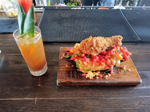 A wooden platter with colorful nachos topped with fried chicken, alongside a tropical drink with a garnish.