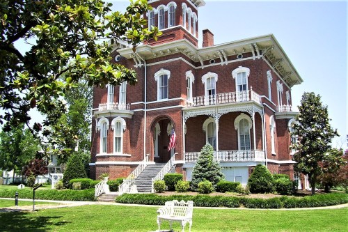 A large, historic brick mansion with multiple balconies, surrounded by lush greenery and a white bench in the foreground.