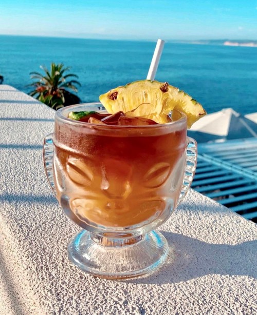 A tropical cocktail in a decorative glass, garnished with a pineapple slice, against a scenic ocean backdrop.