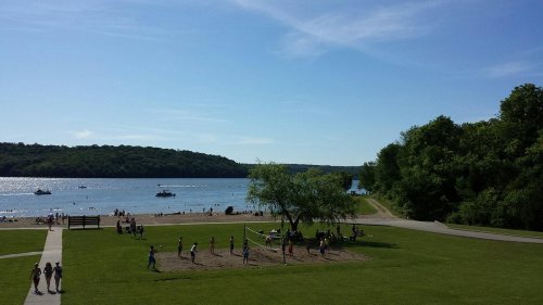A sunny lakeside scene with people playing volleyball on grass, boats on the water, and trees in the background.