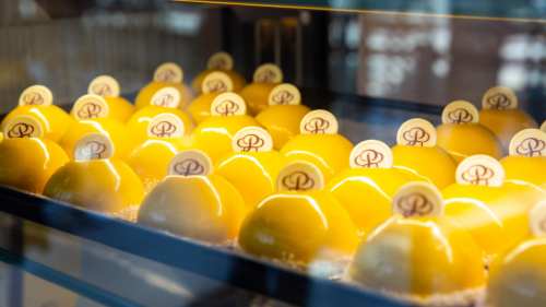 A display of glossy yellow desserts topped with circular logos, arranged neatly in a glass case.