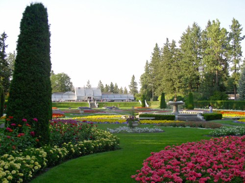 A vibrant garden with colorful flowers, neatly trimmed hedges, and a fountain, surrounded by tall trees and green lawns.