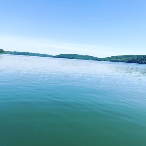 A calm lake reflecting blue skies and distant green hills under bright sunlight.