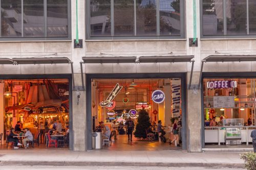 A vibrant market scene with colorful neon signs, outdoor seating, and people dining in a lively urban setting.
