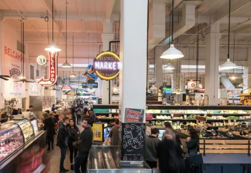 A bustling market interior with various food stalls, neon signs, and shoppers exploring the vibrant space.