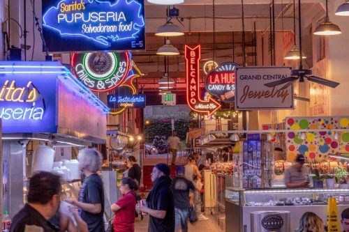 A vibrant market scene with colorful neon signs for various food vendors and shops, bustling with people.