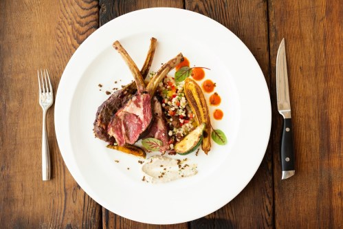A gourmet plate featuring lamb chops, grilled bananas, colorful vegetables, and a creamy sauce on a wooden table.