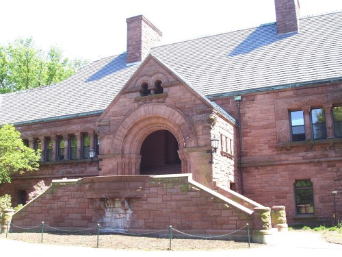 Historic red stone building with a large arched entrance and multiple windows, surrounded by greenery.