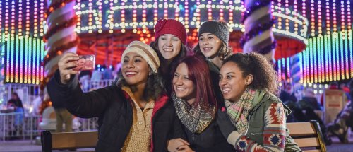 A group of five friends smiling and taking a selfie in front of colorful holiday lights at an amusement park.