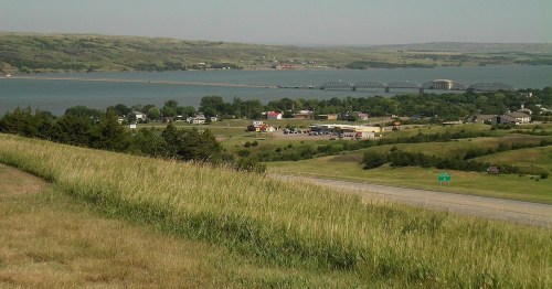 A scenic view of a river, bridge, and small town surrounded by rolling green hills under a clear sky.