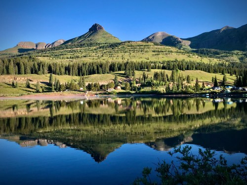 A serene lake reflecting mountains and lush greenery under a clear blue sky.