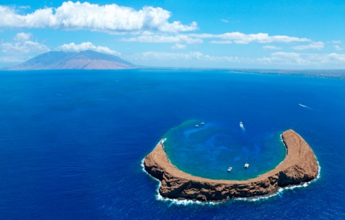 Aerial view of a circular island surrounded by blue ocean, with boats and a distant landmass under a clear sky.