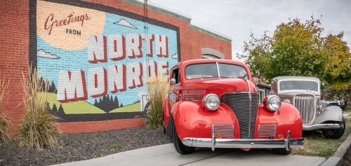 A vintage red car parked in front of a mural that says "Greetings from North Monroe." Another classic car is in the background.