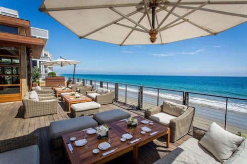 A beachfront patio with cozy seating, tables set for dining, and umbrellas overlooking the ocean and blue sky.