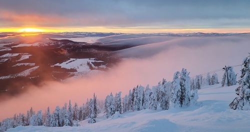 A snowy mountain landscape at sunset, with fog rolling over the valleys and trees dusted in white.