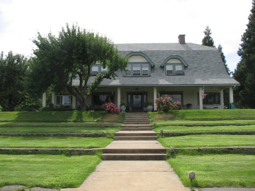 A large, two-story house with a gray roof, surrounded by green lawns and trees, featuring a front porch and steps.