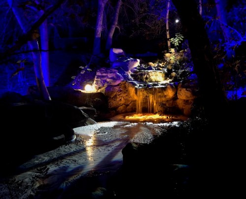 A serene waterfall illuminated by blue lights, surrounded by rocks and trees in a dark, tranquil setting.
