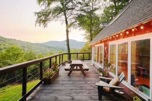 A cozy deck with a picnic table overlooks lush green mountains at sunset, surrounded by trees and soft lighting.