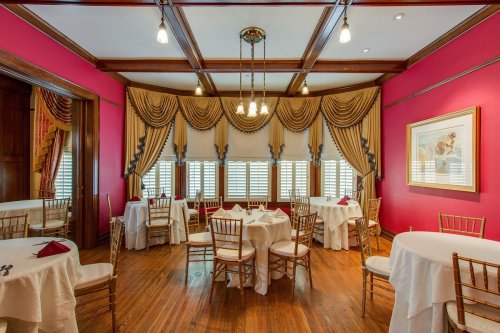 Elegant dining room with pink walls, wooden floors, and tables set with white linens and gold chairs under soft lighting.