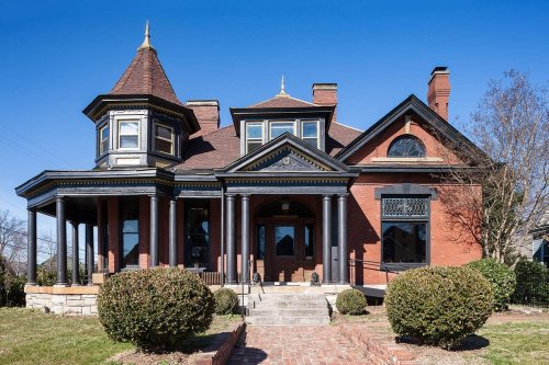 A large, historic brick house with a turret, ornate details, and a landscaped front yard under a clear blue sky.