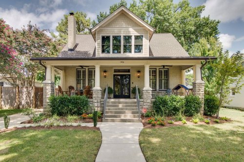 A charming two-story house with a front porch, surrounded by greenery and a well-maintained lawn under a blue sky.