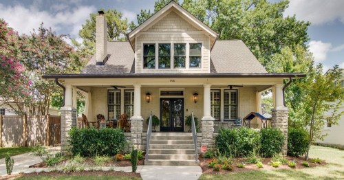 A charming two-story house with a front porch, surrounded by greenery and colorful flowers.