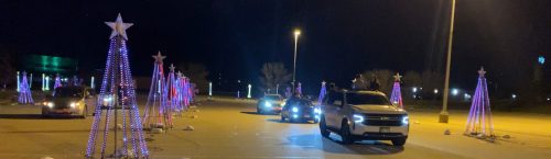 Cars driving through a festive, illuminated display of Christmas trees with stars at night.