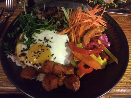 A plate of food featuring rice, a fried egg, sautéed vegetables, and seasoned meat, garnished with herbs.