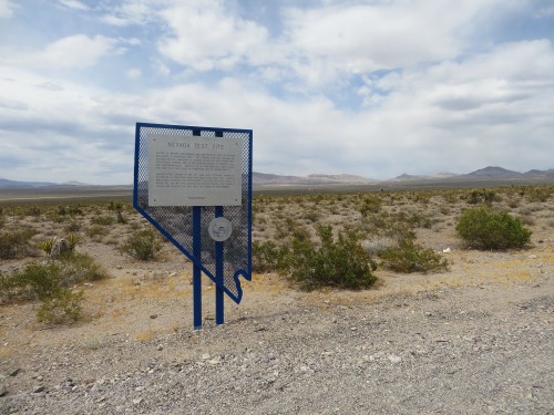 Sign marking the Nevada Test Site, surrounded by desert landscape and mountains under a cloudy sky.