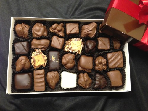 A box of assorted chocolates, featuring various shapes and textures, with a small gift box tied with a red ribbon.