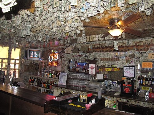 A bar interior with a ceiling covered in dollar bills, a well-stocked bar, and a TV displaying a program.