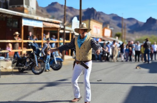 A cowboy in a vest and hat performs in a lively street scene with onlookers and vintage buildings in the background.