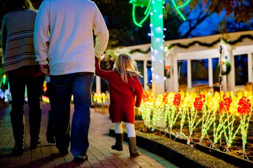 A child in a red dress walks hand-in-hand with an adult, surrounded by colorful holiday lights.