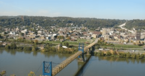 Aerial view of a town with a bridge over a river, surrounded by green hills and buildings under a clear blue sky.