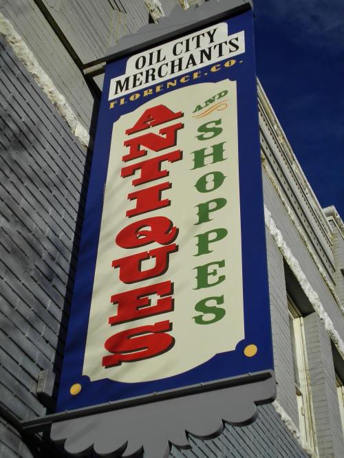 Sign for "Oil City Merchants" featuring "Antiques and Shoppes" in bold, colorful letters against a blue sky.