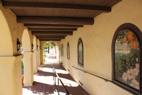 A sunlit corridor with arched windows and a tiled floor, featuring decorative stained glass and wooden beams above.