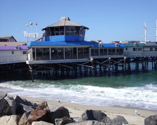 A seaside restaurant on a pier with a blue roof, surrounded by waves and rocks under a clear blue sky.