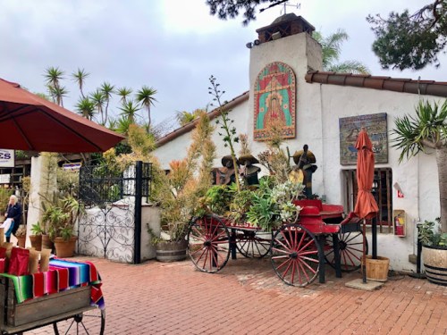 Colorful plants and decorations adorn a rustic cart outside a charming building, with palm trees in the background.