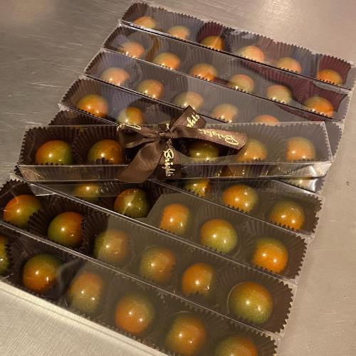 A box of chocolate truffles arranged in rows, each topped with a shiny, colorful coating and tied with a ribbon.