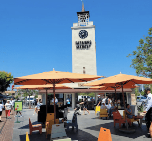 A sunny farmers market scene with orange umbrellas, a clock tower, and people enjoying the outdoor space.
