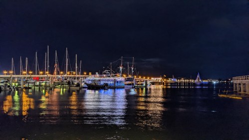 A nighttime view of a marina with illuminated boats and reflections on the water, under a dark sky.