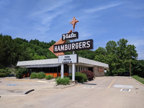 A retro hamburger restaurant with a large sign, surrounded by greenery and a clear blue sky.