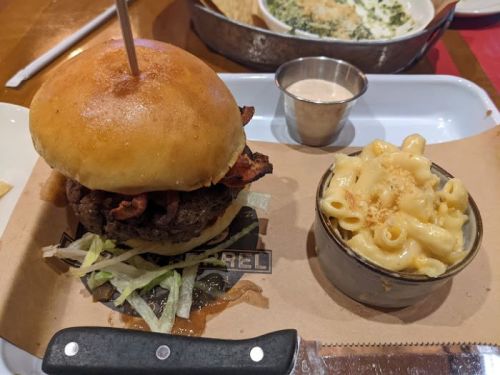A juicy burger with bacon and lettuce on a bun, served with a side of creamy macaroni and cheese in a small bowl.